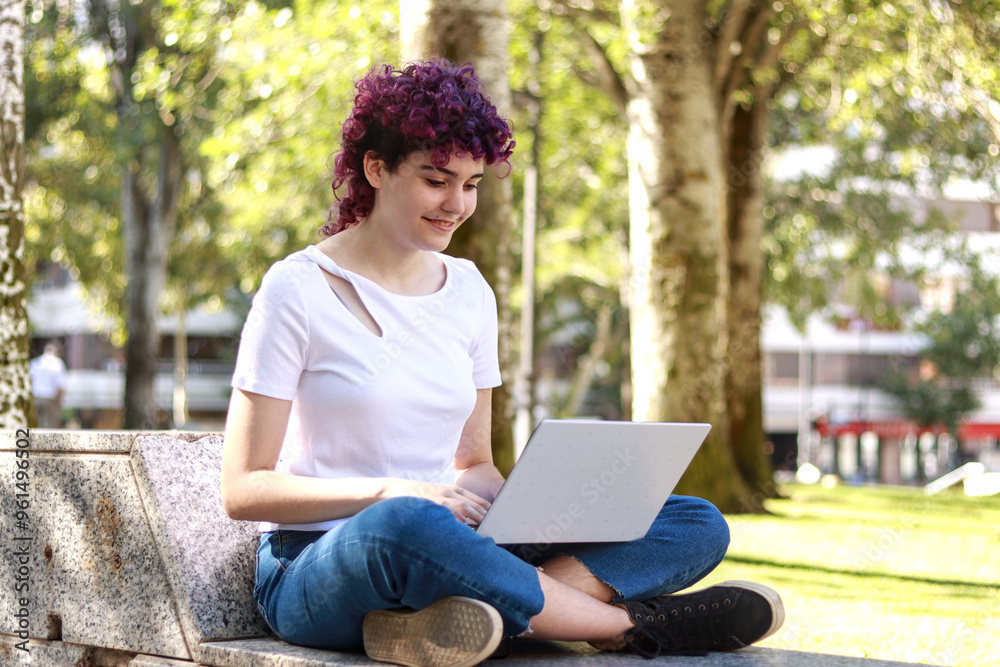 Relaxed young woman with purple curly hair using her laptop on a park bench during a sunny day, surrounded by greenery and natural light Relaxed young woman with purple curly hair using her laptop on a park bench during a sunny day, surrounded by greenery and natural light