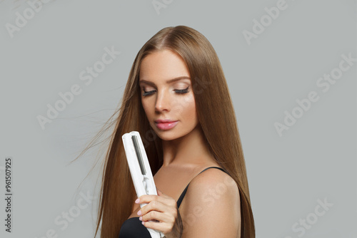 Charming fashion model with brown shiny straight haistyle portrait. Cute woman with straightening iron in her hand on gray studio background photo