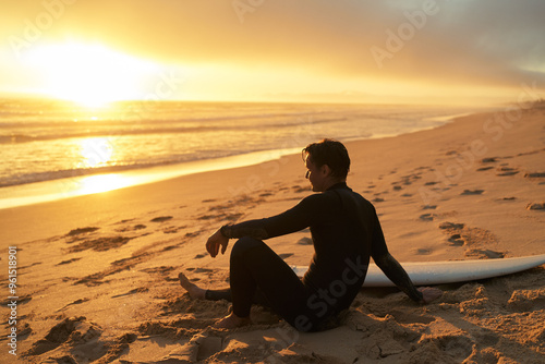 A surfer in wetsuit sits on the sandy beach with a surfboard at golden sunset, reflecting on the serene ocean view. photo