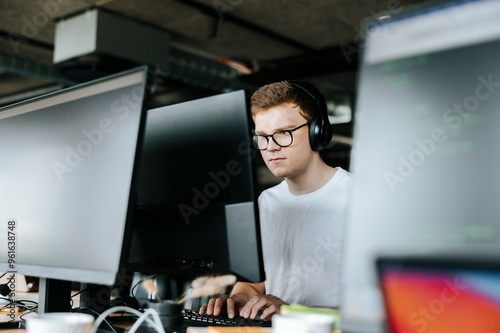 Young male computer programmer typing on desktop PC while sitting on desk in office