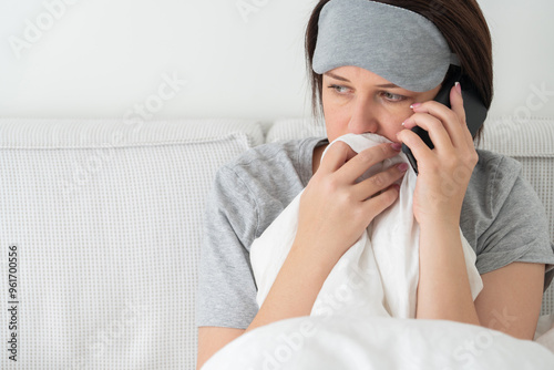 Sad woman wearing sleeping mask using smartphone as she sits in bed covered with duvet