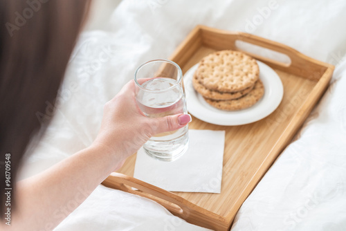 Female hand holding glass of water over tray with crackers on bed