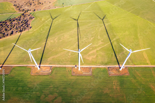 Directly above view of of three towering wind turbines casting long shadows over lush green fields, highlighting the integration of Renewable Energy into a rural landscape with a country road     