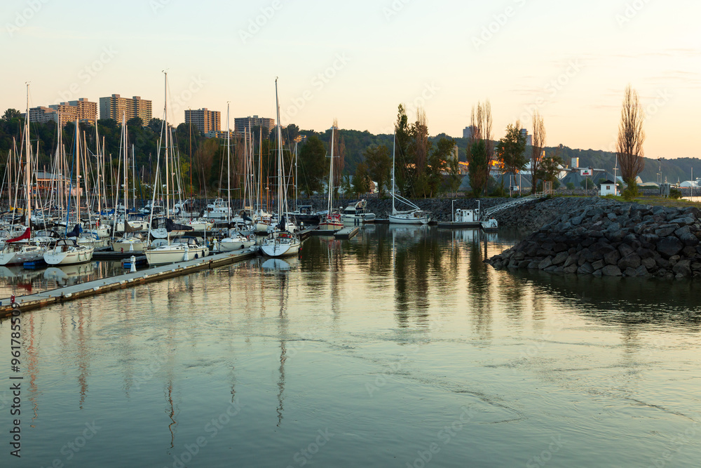 Sailboats moored in the Sillery marina during a golden hour summer morning, with high-rises on cliff in the background, Quebec City, Quebec, Canada Sailboats moored in the Sillery marina during a golden hour summer morning, with high-rises on cliff in the background, Quebec City, Quebec, Canada