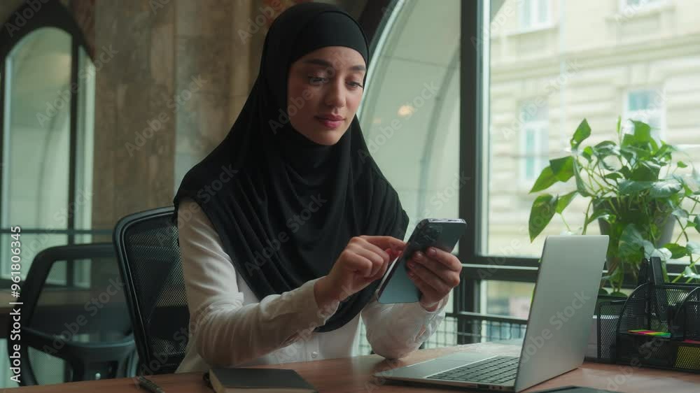 Muslim woman in hijab islamic girl Arabian businesswoman entrepreneur calm smiling business lady working at desk scrolling mobile phone gadget addicted female online app cellphone internet in office Muslim woman in hijab islamic girl Arabian businesswoman entrepreneur calm smiling business lady working at desk scrolling mobile phone gadget addicted female online app cellphone internet in office