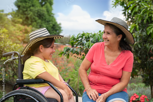 Young Woman in Wheelchair Smiling and Enjoying a Sunny Day in the Countryside with Her Grandmother, Both Wearing Traditional Hats photo