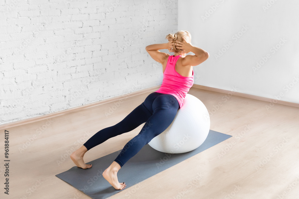 Fit adult woman in pink tank top practice back extension exercise with hands behind her head using fitness ball in loft studio indoor, back view. Fit adult woman in pink tank top practice back extension exercise with hands behind her head using fitness ball in loft studio indoor, back view.