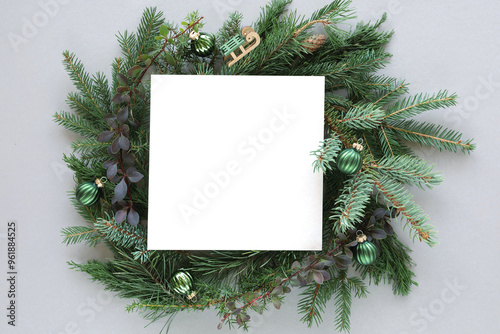 Overhead view of a blank piece of square paper in the middle of a coniferous Christmas wreath decorated with christmas baubles