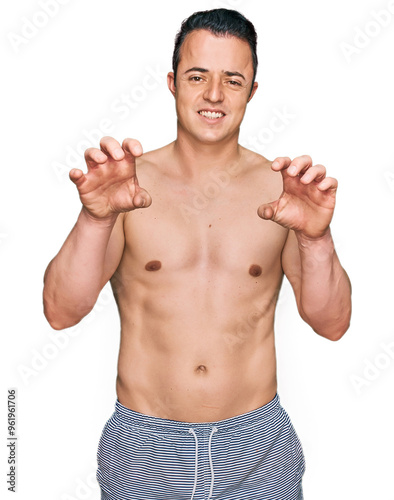 Handsome young man wearing swimwear shirtless smiling funny doing claw gesture as cat, aggressive and sexy expression