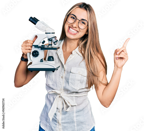 Hispanic young woman holding microscope smiling happy pointing with hand and finger to the side