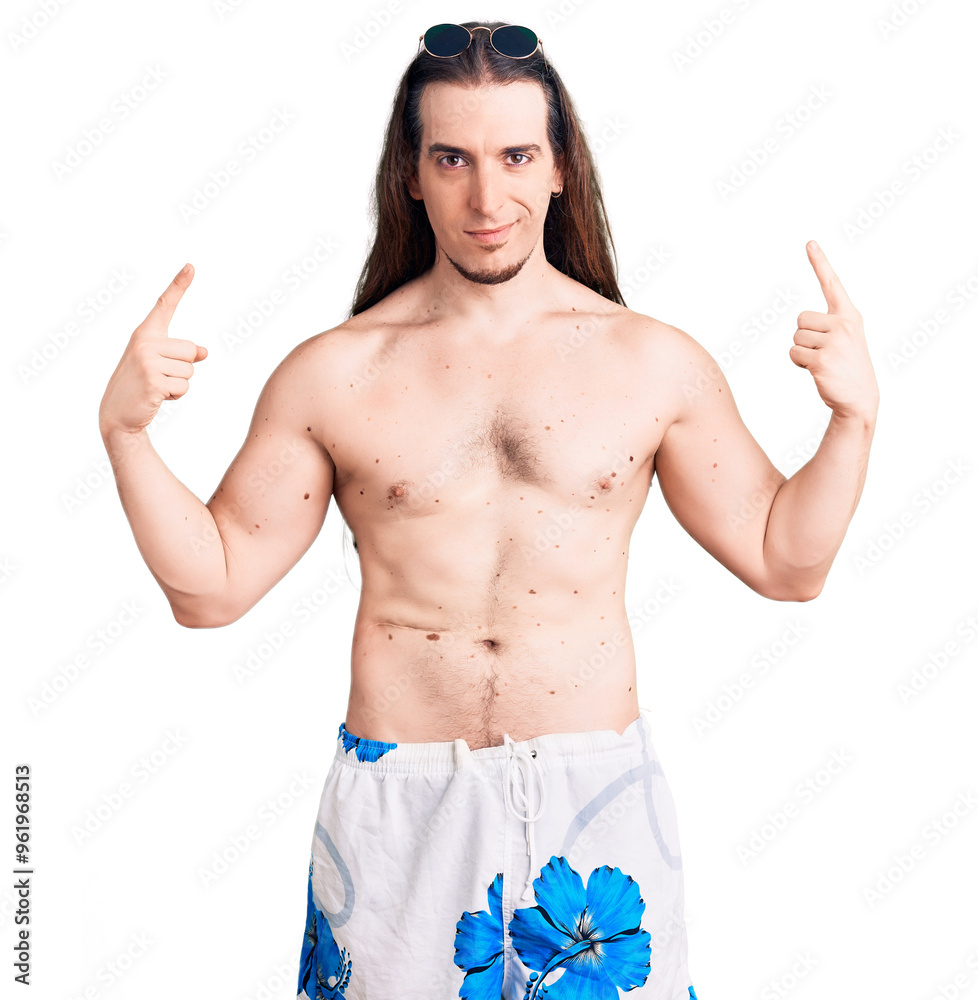 Fototapeta premium Young adult man with long hair wearing swimwear shirtless looking confident with smile on face, pointing oneself with fingers proud and happy.