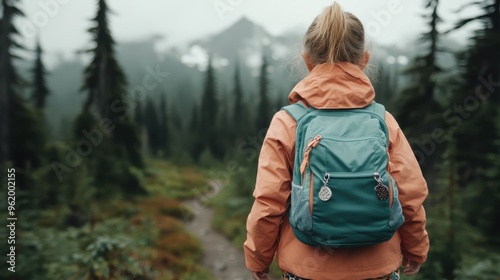 A young hiker with a teal backpack is seen from behind, standing on a forest path with a picturesque mountain view in the background, capturing the essence of adventure and nature. photo