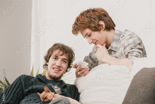 Two male friends relaxing on sofa with dog and looking at smartp photo