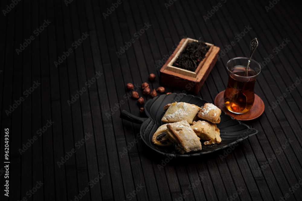 Minimalistic scene with traditional pastries on a black plate, hazelnuts, and a glass of tea. A rustic wooden box with loose tea leaves enhances the cozy, authentic tea-time atmosphere. Minimalistic scene with traditional pastries on a black plate, hazelnuts, and a glass of tea. A rustic wooden box with loose tea leaves enhances the cozy, authentic tea-time atmosphere.