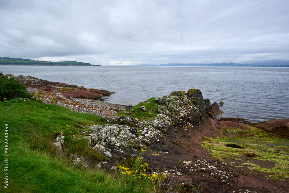 A serene rocky shore with lush grass, flowers, and tranquil water. A solitary figure contemplates the vast scenery. Details include rugged textures and vibrant yellow flowers. A serene rocky shore with lush grass, flowers, and tranquil water. A solitary figure contemplates the vast scenery. Details include rugged textures and vibrant yellow flowers.