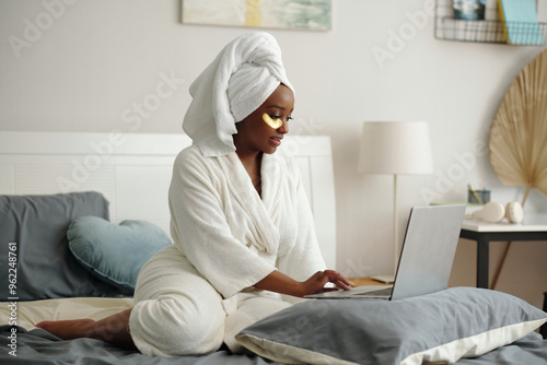 Person in white bathrobe and head wrap sitting on bed, applying skincare routine. Bedroom features subtle decor, with soft lighting emanating from a lamp in the corner