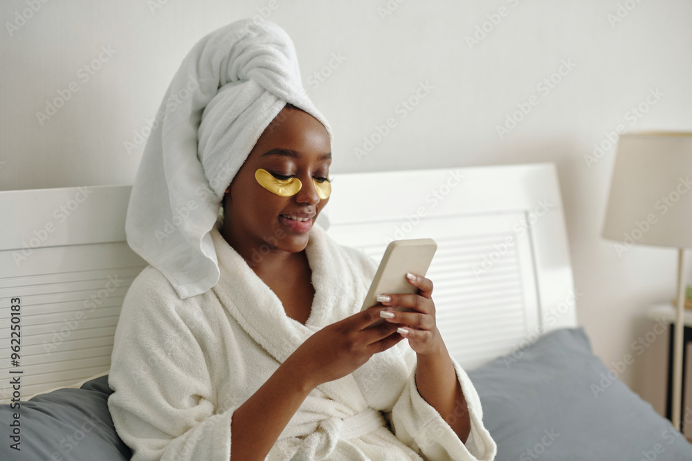 Woman wearing bathrobe and head wrap sitting on bed, applying under-eye patches while holding smartphone and smiling with bedroom lamp in background and looking peaceful Woman wearing bathrobe and head wrap sitting on bed, applying under-eye patches while holding smartphone and smiling with bedroom lamp in background and looking peaceful