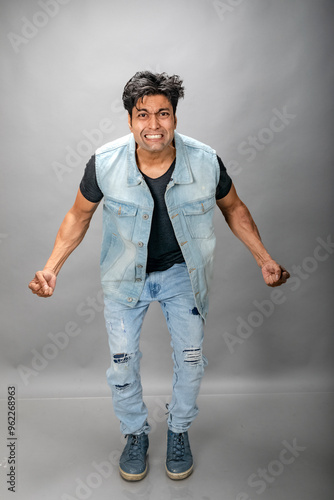 Angry man yelling and shaking hands, grimacing with hatred, standing hateful against grey background wearing denim jeans and black t-shirt