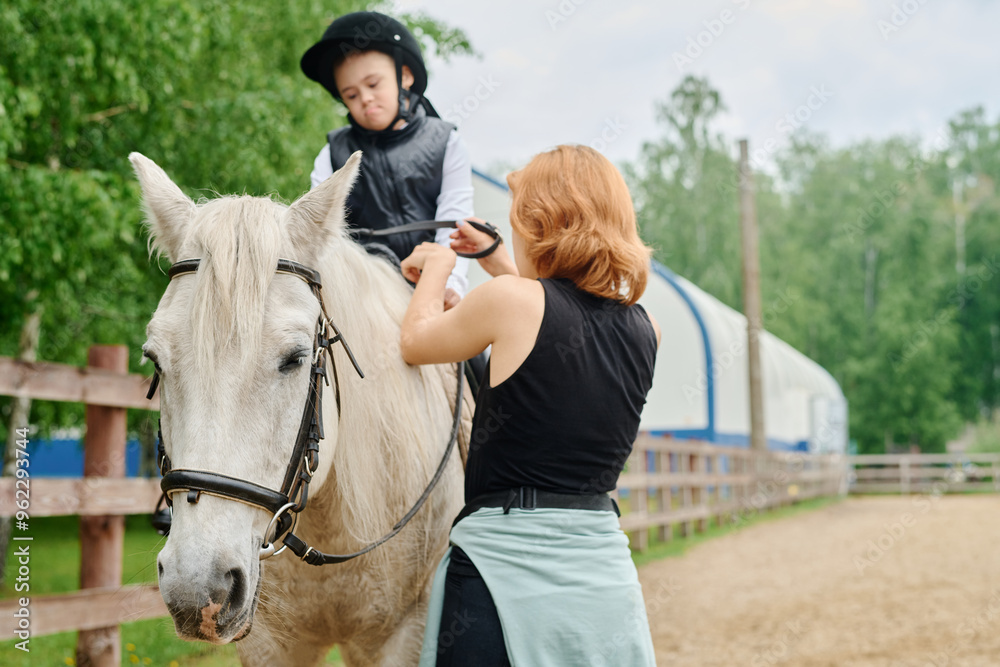 Child riding a white horse being assisted by an adult instructor on an outdoor riding arena with greenery in background Child riding a white horse being assisted by an adult instructor on an outdoor riding arena with greenery in background