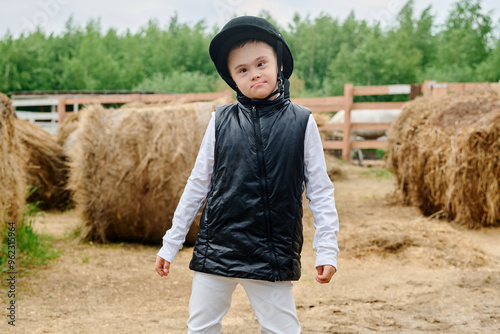 Young child with Down syndrome wearing equestrian helmet posing in farm environment with haystacks and greenery around creating serene rural backdrop