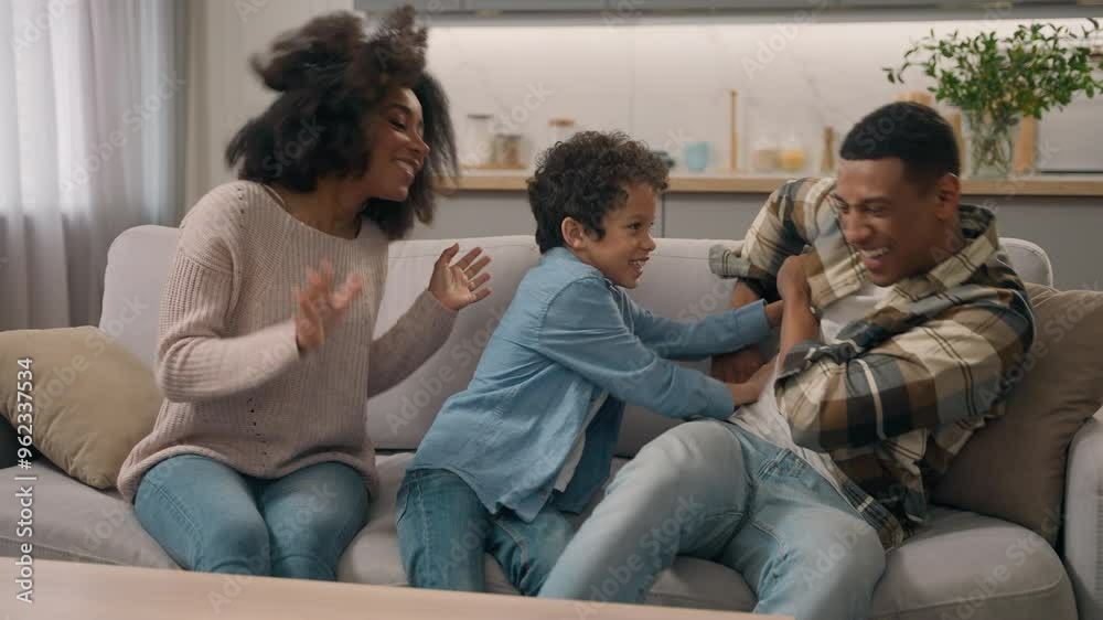 Happy family having fun African American joyful couple female mom woman male dad man cuddle hug embrace playing tickling their adopted child little boy laughing son kid inside modern living room flat Happy family having fun African American joyful couple female mom woman male dad man cuddle hug embrace playing tickling their adopted child little boy laughing son kid inside modern living room flat