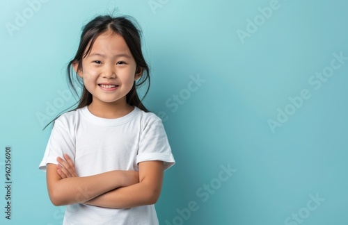 A smiling young girl with dark hair and a white shirt stands against a teal background with her arms crossed. Her bright eyes and happy expression convey a sense of joy and charm.