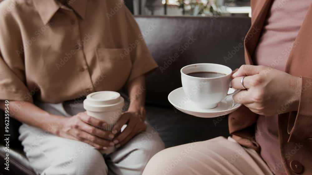 Cropped shot of unrecognizable businesswomen in smart casual clothes having nice cup of coffee during break while sitting on leather couch and communicating in office