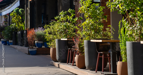A restaurant features an inviting outdoor seating area lined with vibrant green plants and flowers on a bright, sunny day
