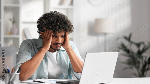 A young Indian male businessman is sitting at a desk trying to work, checking schedules and getting frustrated that something is not working out. Headache, fatigue, burnout.