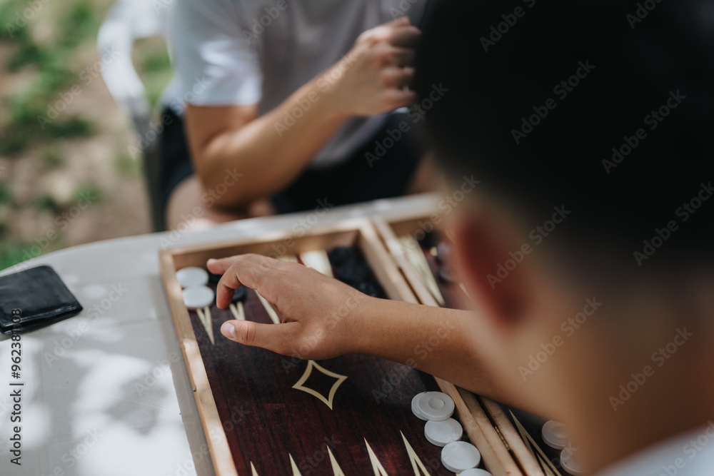 Close-up of two people playing backgammon at a table outdoors, capturing a moment of leisure and strategy. Close-up of two people playing backgammon at a table outdoors, capturing a moment of leisure and strategy.