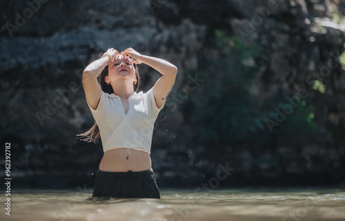 A young woman stands waist-deep in a serene natural body of water, basking in sunlight while enjoying a refreshing moment.