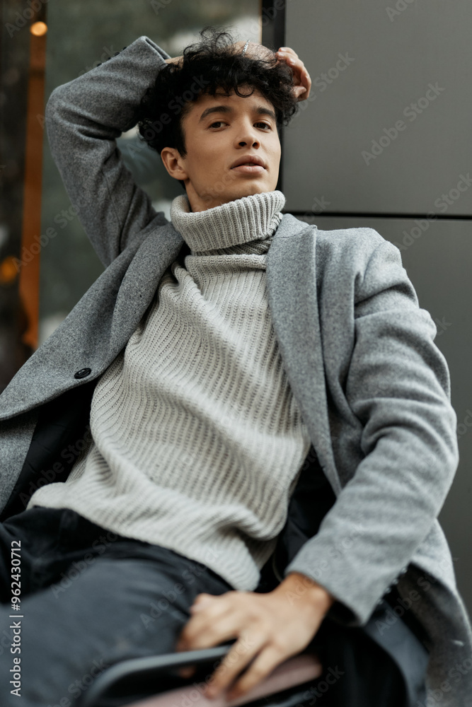 Fashionable young man in gray sweater and black pants sitting on a chair with hands on head, looking at camera Fashionable young man in gray sweater and black pants sitting on a chair with hands on head, looking at camera