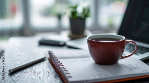 A red cup of tea on a saucer sits on a desk next to an open notebook and a pen. The organized workspace is perfect for focused work or study, with a plant adding a touch of nature.