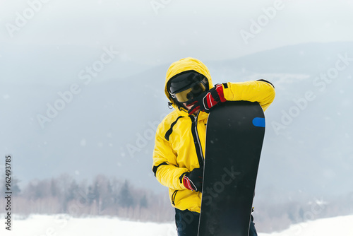 Young guy snowboarder looks mysteriously at the camera. Holds a snowboard in his hands against the background of mountains. Active recreation, snowboarding at resort 