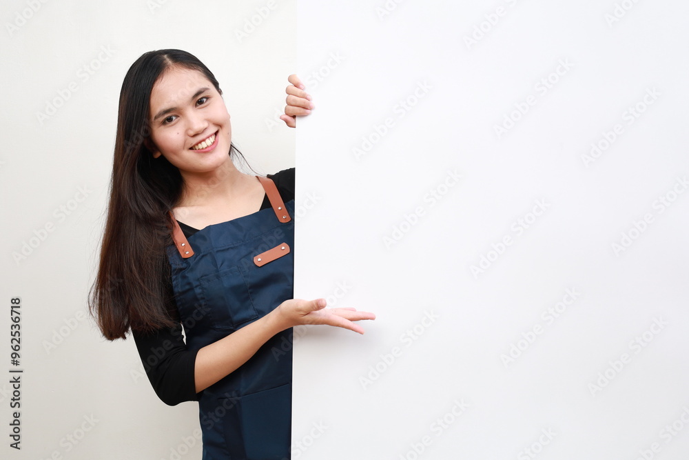 Cheerful Asian woman in apron presenting something on white advertisement board on white background Cheerful Asian woman in apron presenting something on white advertisement board on white background