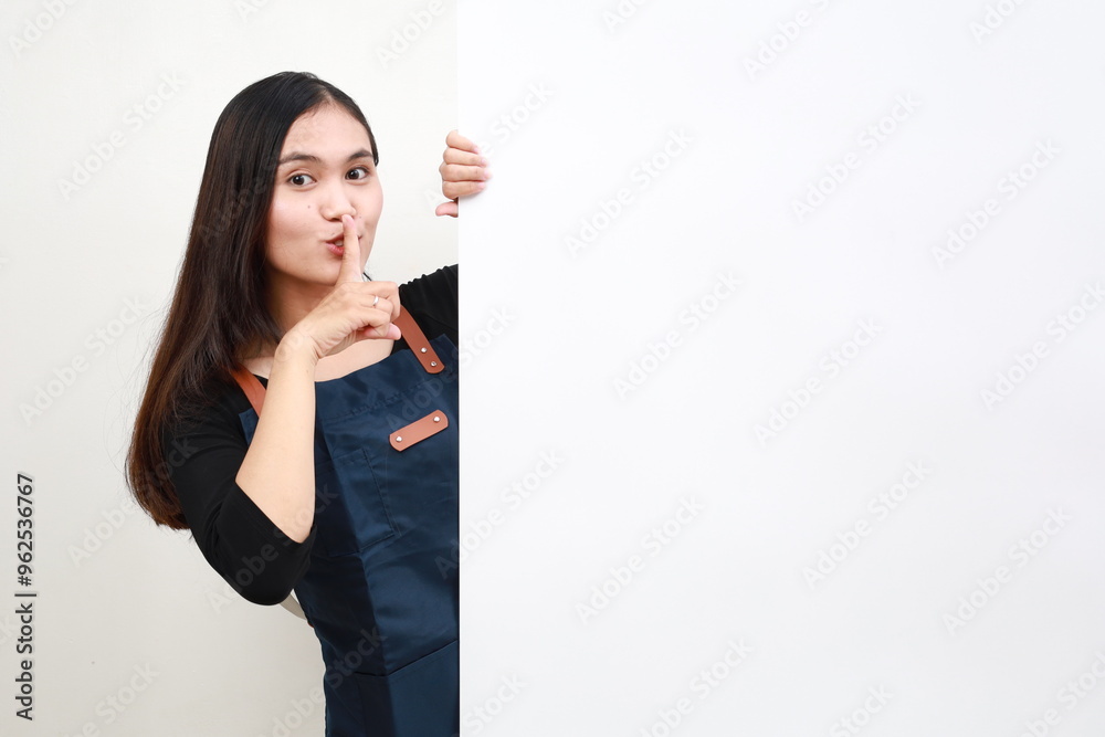 Smiling Asian woman in blue apron peeking behind wall with silent gesture Smiling Asian woman in blue apron peeking behind wall with silent gesture