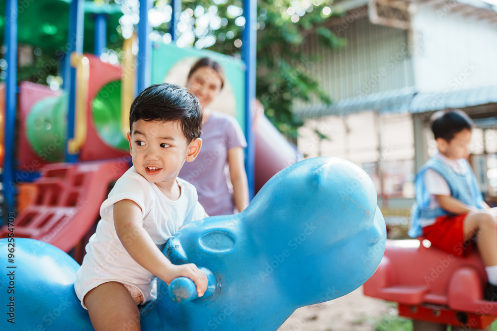 Obraz premium A young Asian boy, happy playing as rides a rocking horse at the playground, his mother standing close by to watch over him and ensure his safety during playtime.