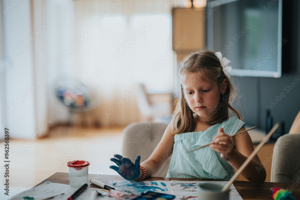 Fototapeta premium A focused young girl paints with watercolors at home, expressing creativity and concentration during an art activity.