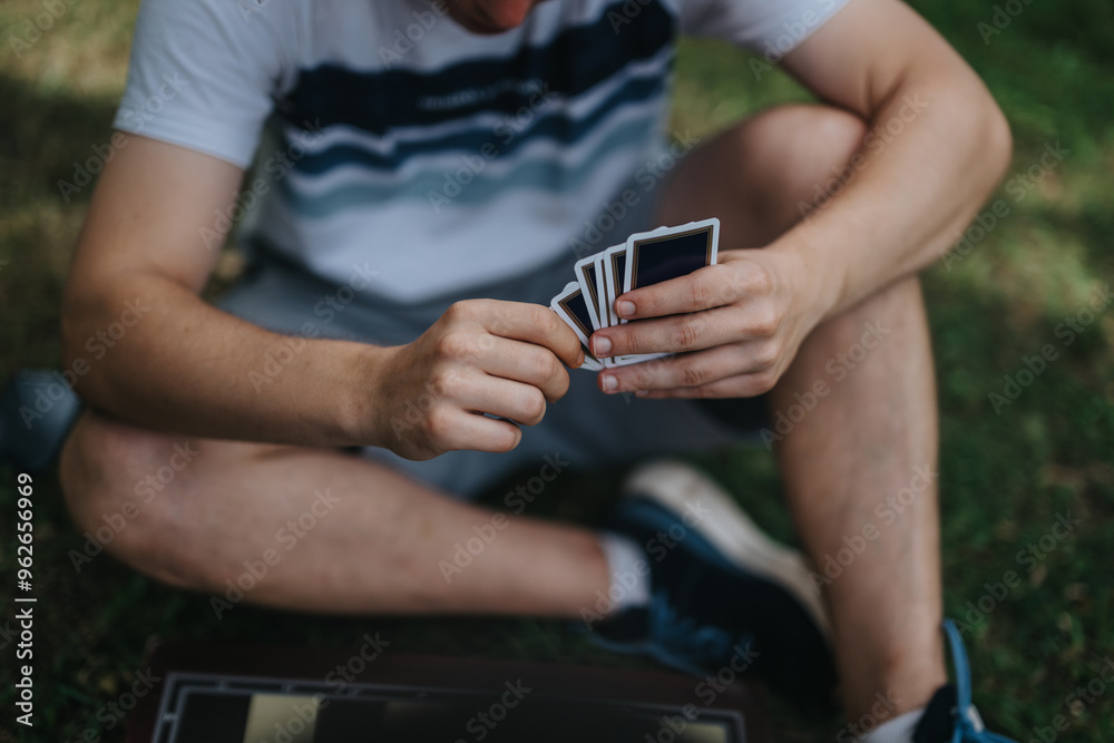 An individual sits cross-legged on grass, holding playing cards during an outdoor game, enjoying leisure time. An individual sits cross-legged on grass, holding playing cards during an outdoor game, enjoying leisure time.