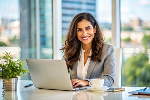 A Confident Hispanic Businesswoman Smiling And Working On A Laptop At A Desk In A Brightly Lit Office