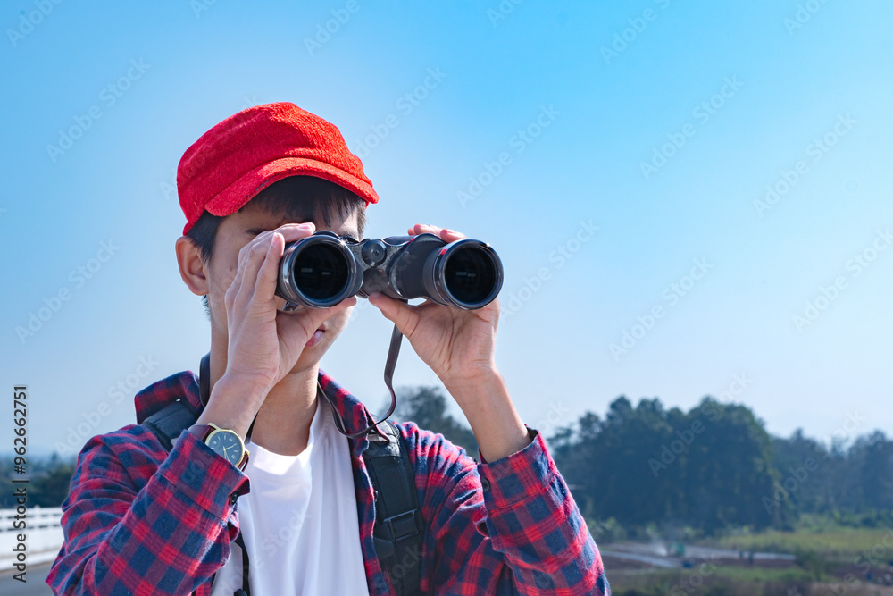 Obraz premium Asian boy wearing a red cap and plaid shirt, looking through binoculars while standing on a bridge over a river. He’s enjoying the natural surroundings and observing wildlife with curiosity.