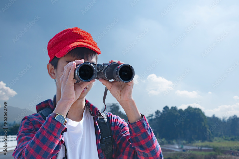 Obraz premium Asian boy wearing a red cap and plaid shirt, looking through binoculars while standing on a bridge over a river. He’s enjoying the natural surroundings and observing wildlife with curiosity.