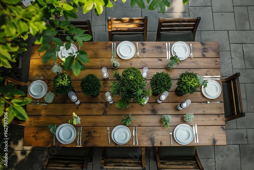 Outdoor dining setup on wooden table with white tablecloth, candles, green plants. Mini topiary centerpieces surround tree and bush. Perfect for garden party outdoor wedding decoration. photo