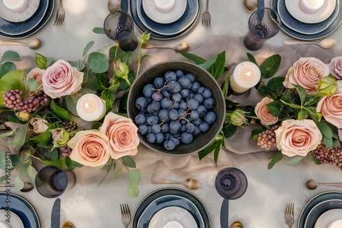 Formal dinner table setting with white tablecloth, blue runner, black bowl centerpiece, dark purple grapes, pink and white flowers. Elegant atmosphere with gold plates, silverware, and cups. photo