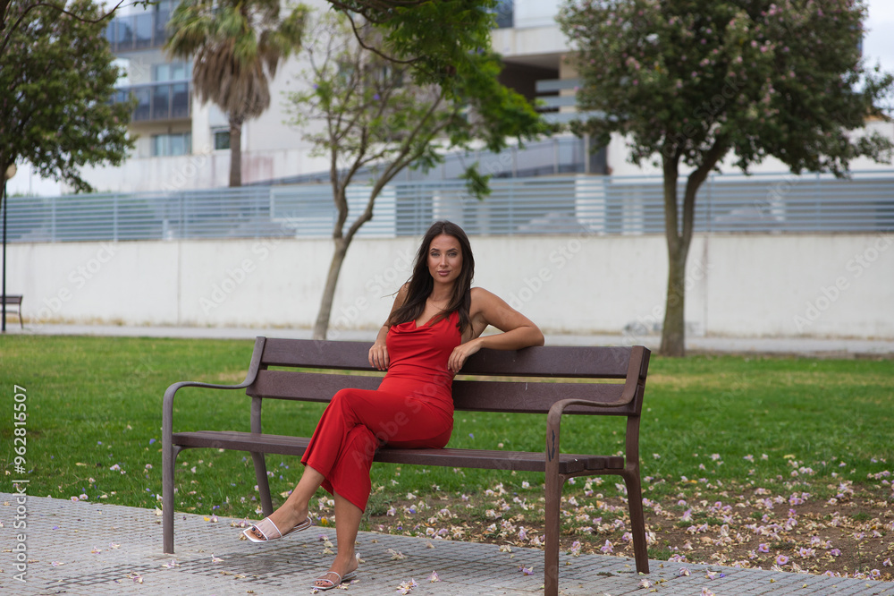 Beautiful young Spanish woman with long hair and green eyes wearing a very elegant red dress sitting on a wooden bench in the park. The woman has her legs crossed and is resting peacefully. Beautiful young Spanish woman with long hair and green eyes wearing a very elegant red dress sitting on a wooden bench in the park. The woman has her legs crossed and is resting peacefully.
