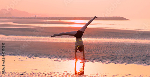 Fotografie young girl in backlight doing a cartwheel with her legs up at the seaside in sum
