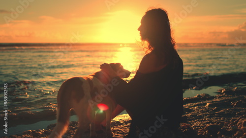Fotografie Woman sitting on beach with her dog enjoy bright colorful orange sunset over the sea