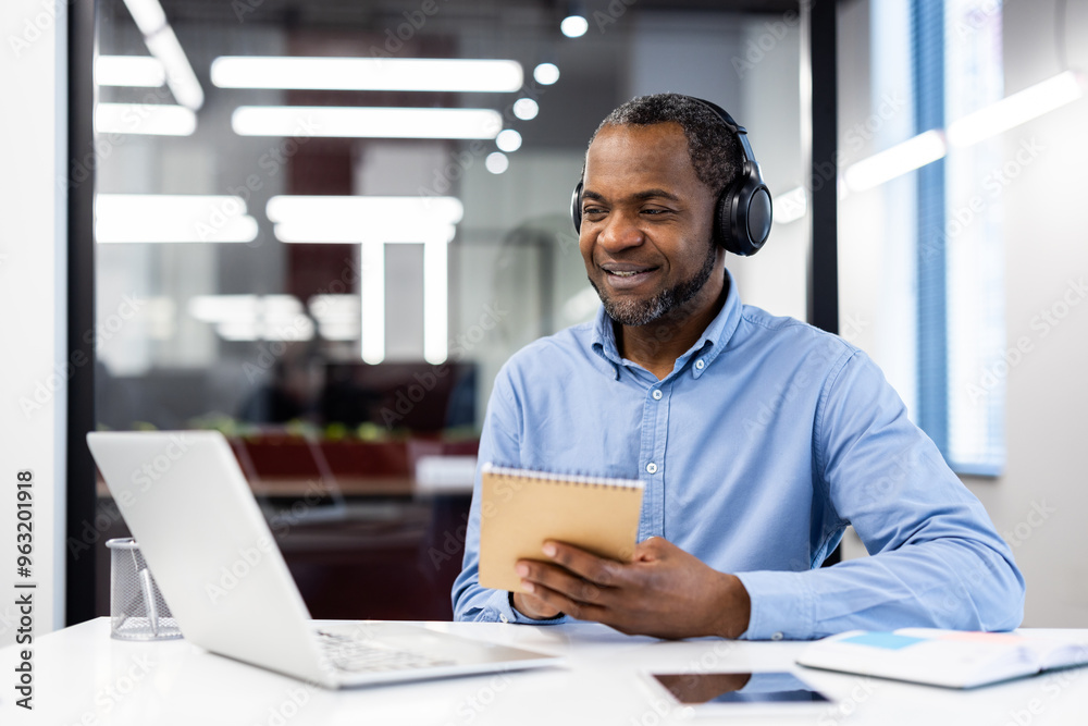 Businessman in blue shirt and headphones studies by watching webinar on laptop in office. He takes notes in notepad, demonstrating focus and engagement in professional development. Businessman in blue shirt and headphones studies by watching webinar on laptop in office. He takes notes in notepad, demonstrating focus and engagement in professional development.