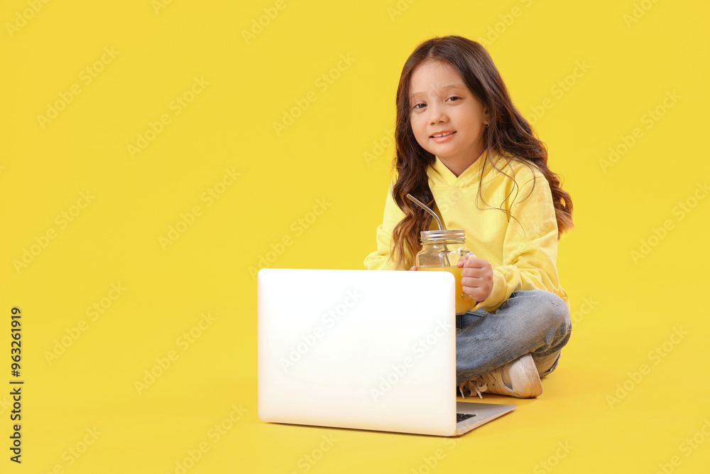 Little Asian girl with mason jar of fresh citrus juice and laptop sitting on yellow background Little Asian girl with mason jar of fresh citrus juice and laptop sitting on yellow background