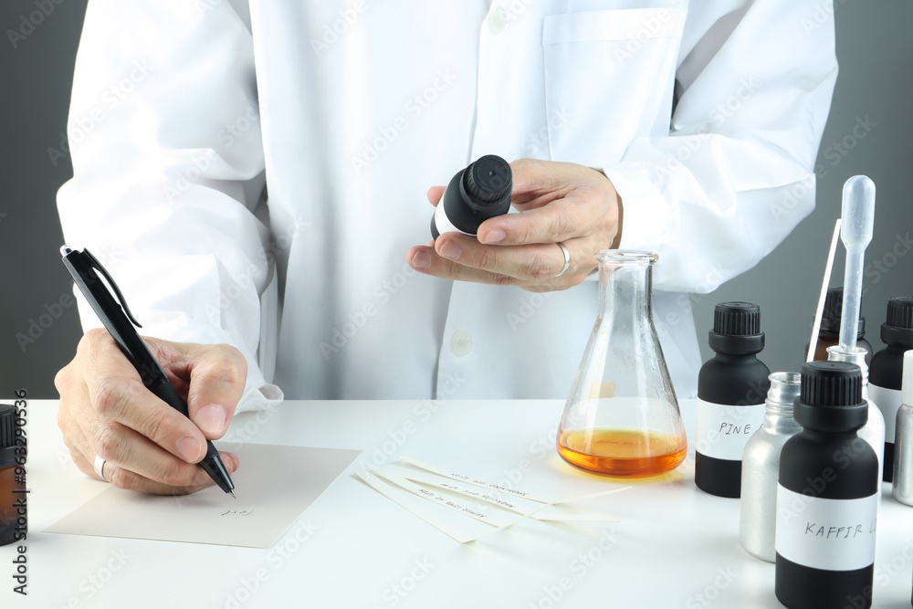 right hand is writing testing result of blending scents for making perfume and candle by perfumer in laboratory with chemical beaker blotting paper , fragrance bottle and essential oil bottle on table right hand is writing testing result of blending scents for making perfume and candle by perfumer in laboratory with chemical beaker blotting paper , fragrance bottle and essential oil bottle on table